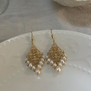 Peut inclure: Une paire de boucles d'oreilles pendantes dor&eacute;es avec une section sup&eacute;rieure en m&eacute;tal ajour&eacute; en forme de losange. Chaque boucle est orn&eacute;e de plusieurs petites perles d'eau douce blanches en forme de goutte suspendues &agrave; la base.