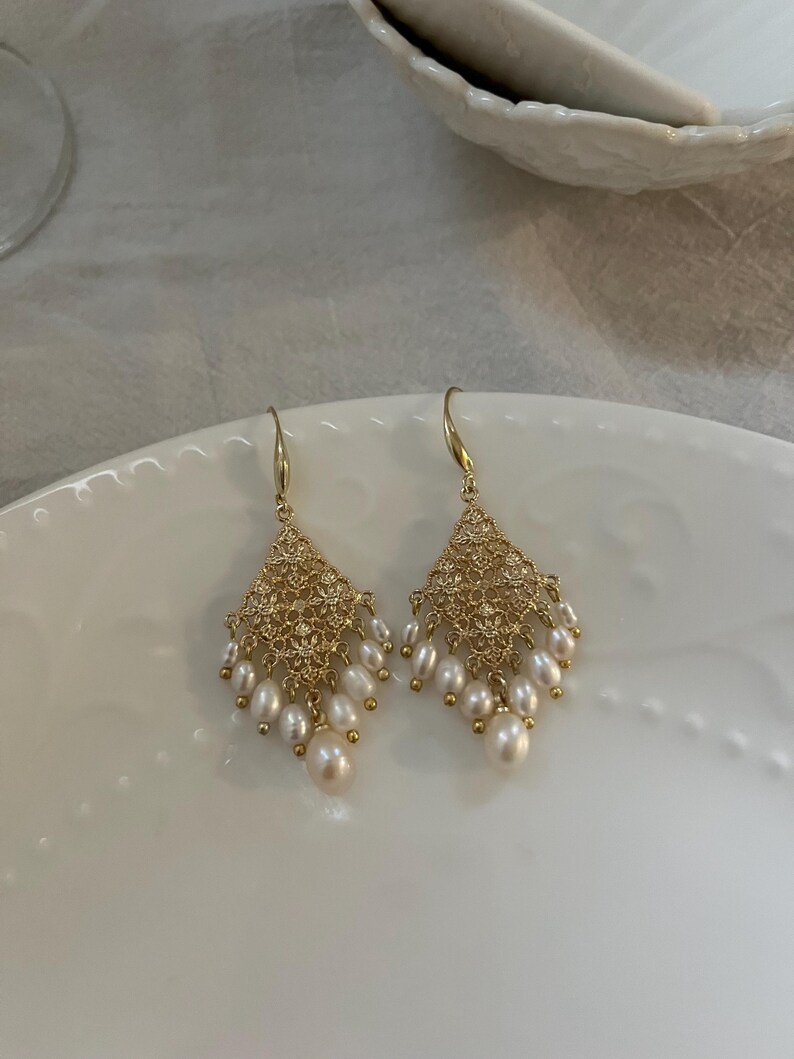 Peut inclure: Une paire de boucles d'oreilles pendantes dor&eacute;es avec une section sup&eacute;rieure en m&eacute;tal ajour&eacute; en forme de losange. Chaque boucle est orn&eacute;e de plusieurs petites perles d'eau douce blanches en forme de goutte suspendues &agrave; la base.