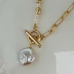 May include: A gold necklace with a large, irregular pearl pendant. The necklace has a toggle clasp and a chain with small beads and larger gold links. The pearl is white with subtle pink and brown hues.