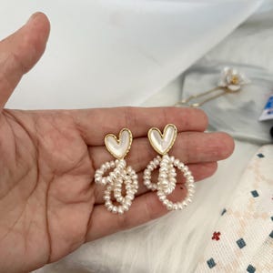 May include: A pair of gold heart-shaped earrings with white pearl accents. The heart is made of a white shell-like material. The pearls are strung in a circle shape.