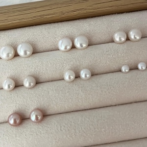 Op de afbeelding: Een verzameling parelstud-oorbellen in verschillende maten en kleuren. De oorbellen zijn gerangschikt op een lichtbeige oppervlak. De parels variëren in kleur van wit tot een zachtroze tint, wat hun natuurlijke glans en ronde vorm benadrukt.