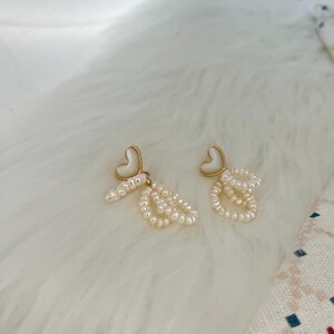 May include: A pair of gold heart-shaped earrings with white pearl accents. The earrings feature a delicate chain of pearls dangling from each heart.