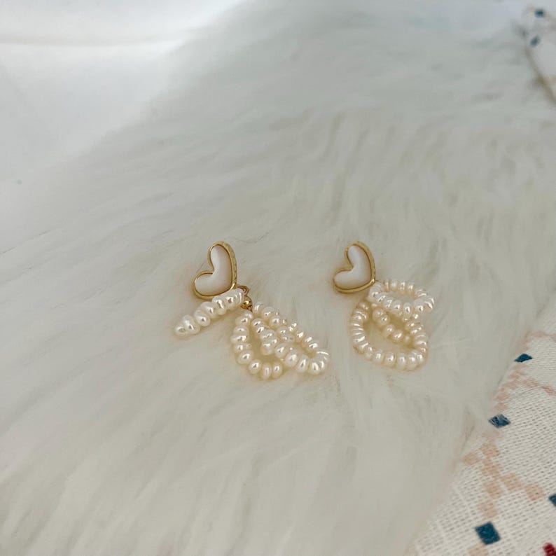 May include: A pair of gold heart-shaped earrings with white pearl accents. The earrings feature a delicate chain of pearls dangling from each heart.