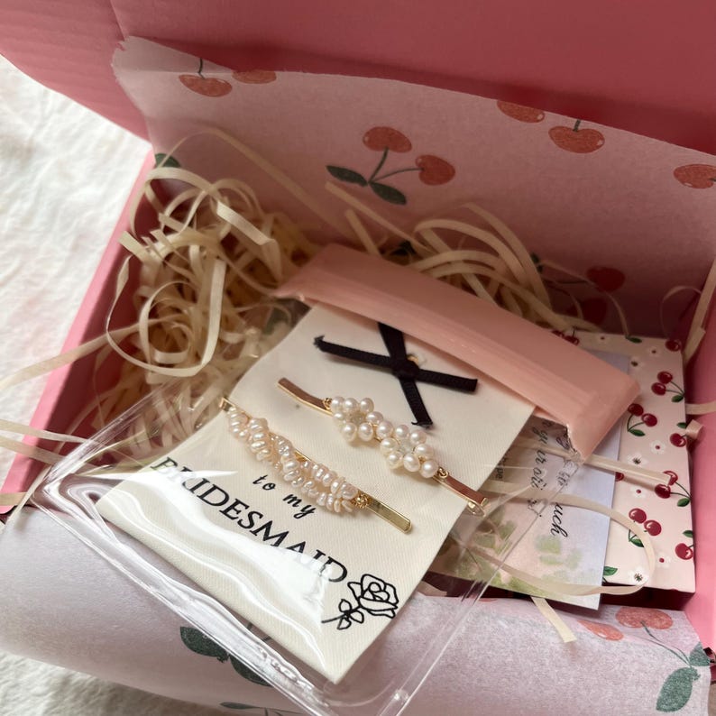 Puede incluir: Una caja de regalo rosa con estampado de cerezas contiene una bolsa transparente con dos pinzas para el pelo de perlas, una pinza para el pelo rosa y una tarjeta peque&ntilde;a que dice "to my BRIDESMAID". La caja est&aacute; llena de virutas de papel.