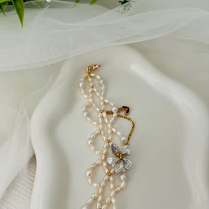 May include: A delicate pearl necklace with a floral pendant. The necklace features multiple strands of small, off-white pearls, accented with gold-colored beads. The pendant is a silver-colored flower with a clear center. The necklace is displayed on a white, curved surface.