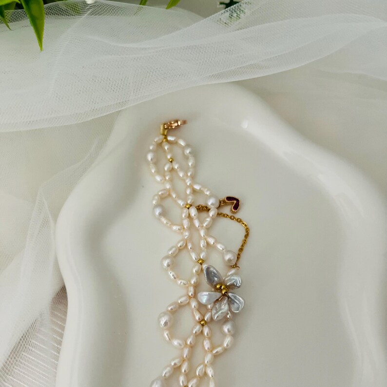 May include: A delicate pearl necklace with a floral pendant. The necklace features multiple strands of small, off-white pearls, accented with gold-colored beads. The pendant is a silver-colored flower with a clear center. The necklace is displayed on a white, curved surface.