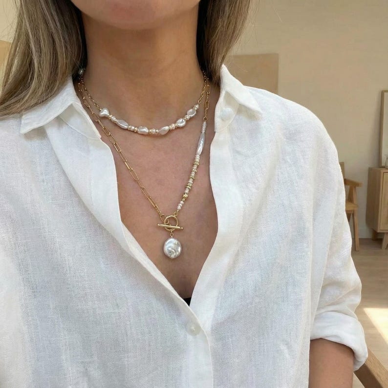May include: A layered necklace set with gold chains and natural pearls. The top layer has a strand of small pearls, while the lower layer includes a toggle clasp and a large, irregular pearl pendant. The jewelry is worn over a white button-down shirt.