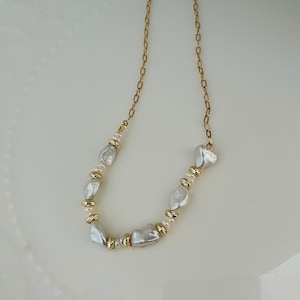 May include: A gold-coloured chain necklace adorned with irregular white pearls and gold-toned beads. The necklace's design incorporates a series of pearls and gold accents. The pearls have an organic shape, adding to the necklace's elegant and unique design.