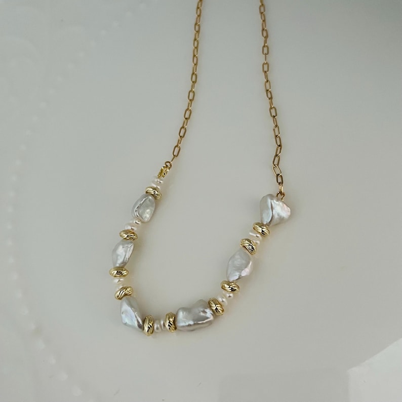 May include: A gold-coloured chain necklace adorned with irregular white pearls and gold-toned beads. The necklace's design incorporates a series of pearls and gold accents. The pearls have an organic shape, adding to the necklace's elegant and unique design.