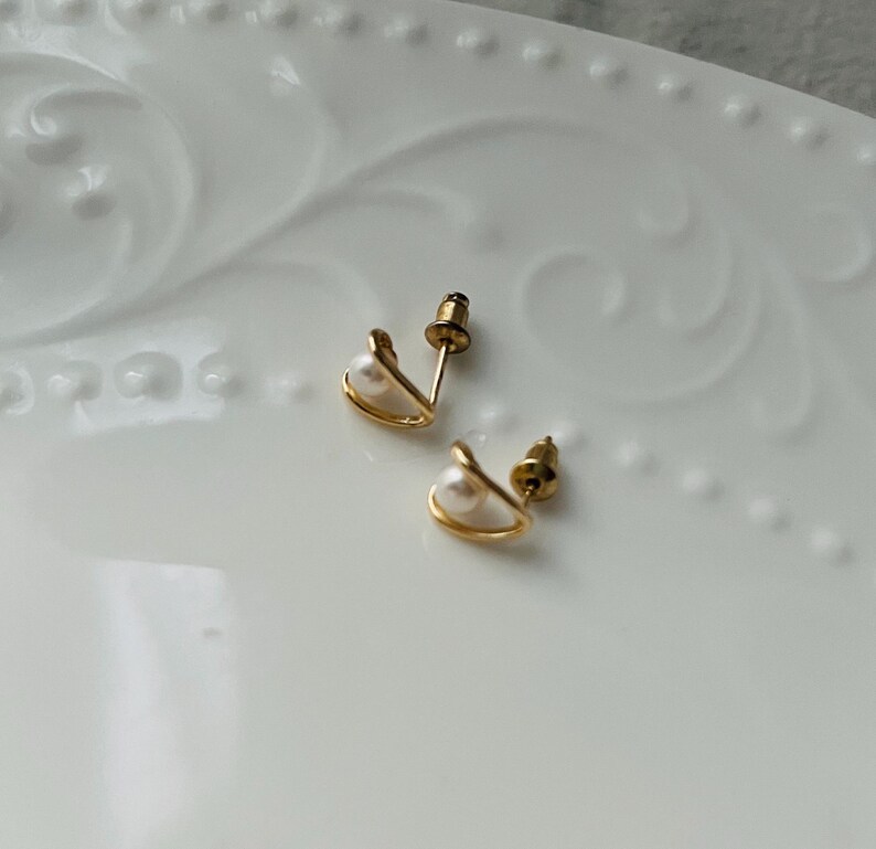 K&ouml;nnte beinhalten: Ein Paar goldene Ohrringe mit einer kleinen Perle in jedem. Die Ohrringe haben die Form einer geschwungenen Linie.