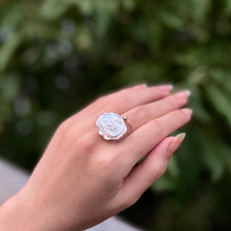 May include: A white pearl ring with a flower design. The ring is on a person's finger.
