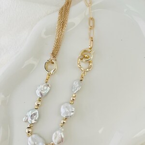Peut inclure: Un collier dor&eacute; orn&eacute; de perles blanches irr&eacute;guli&egrave;res et de petites perles dor&eacute;es. Le collier pr&eacute;sente une cha&icirc;ne &agrave; plusieurs brins d'un c&ocirc;t&eacute; et un motif de maillons de cha&icirc;ne de l'autre, avec un fermoir d&eacute;coratif.