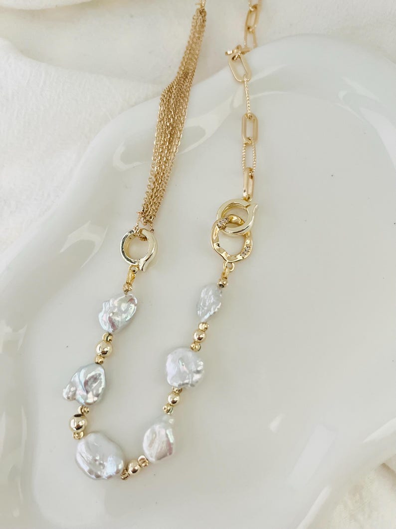 Peut inclure: Un collier dor&eacute; orn&eacute; de perles blanches irr&eacute;guli&egrave;res et de petites perles dor&eacute;es. Le collier pr&eacute;sente une cha&icirc;ne &agrave; plusieurs brins d'un c&ocirc;t&eacute; et un motif de maillons de cha&icirc;ne de l'autre, avec un fermoir d&eacute;coratif.