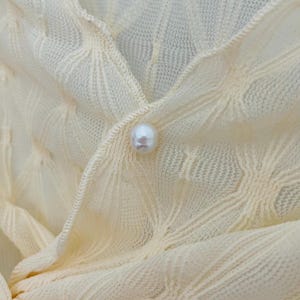 May include: A close-up of a cream-colored knitted cardigan with a pearl button.