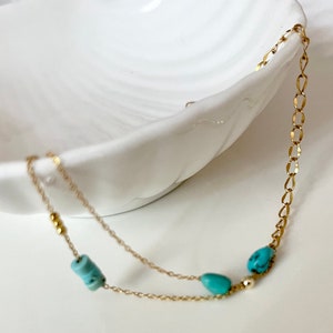 May include: A delicate gold chain necklace with turquoise beads. The necklace is layered with two chains and features small gold beads.