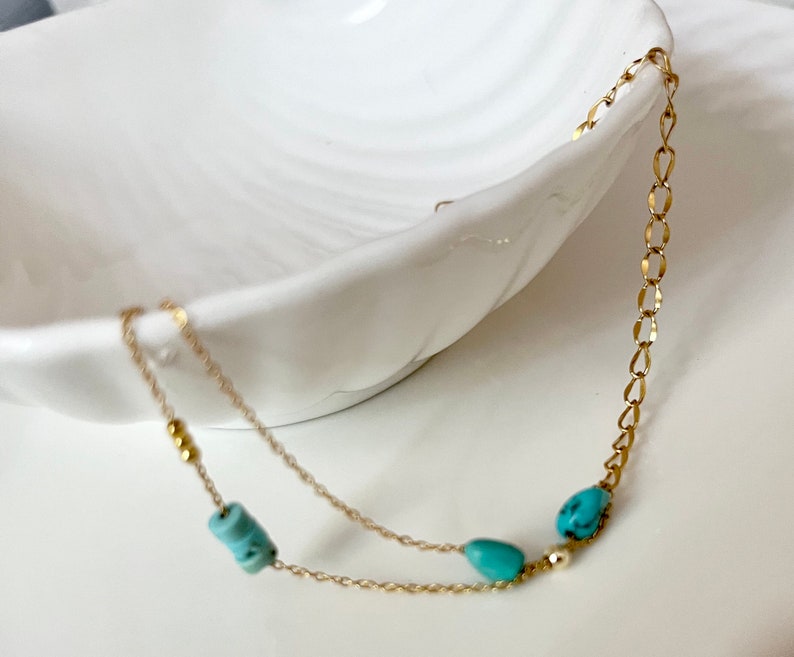 May include: A delicate gold chain necklace with turquoise beads. The necklace is layered with two chains and features small gold beads.