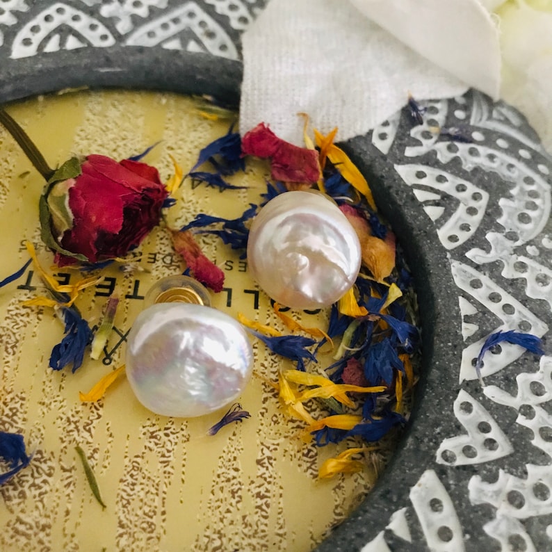 May include: A pair of white pearl earrings with gold posts. The pearls are round and have an iridescent sheen. The earrings are displayed on a background of dried flowers.