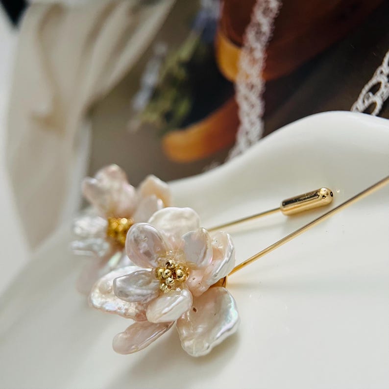 Puede incluir: Un broche de flor de perla delicado con un alfiler de oro. La flor est&aacute; hecha de perlas blancas y rosadas y tiene un centro de oro.