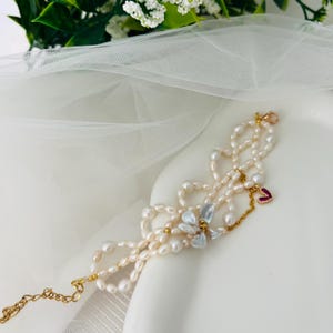 May include: A delicate pearl bracelet with multiple strands, featuring a gold chain and clasp. The bracelet is adorned with a small red heart charm and a cluster of iridescent, petal-shaped beads. The pearls are off-white and the metal is gold.