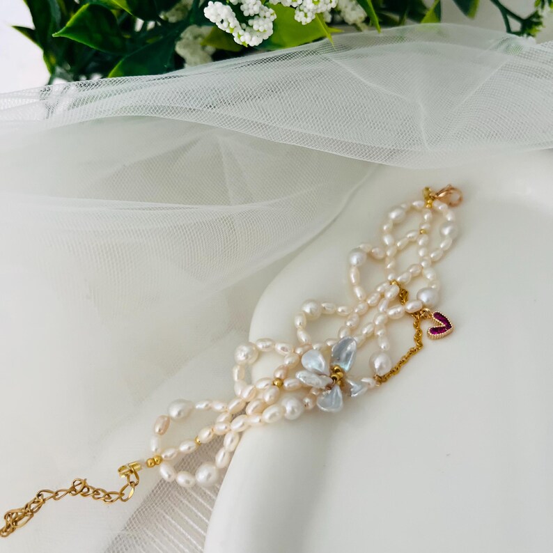 May include: A delicate pearl bracelet with multiple strands, featuring a gold chain and clasp. The bracelet is adorned with a small red heart charm and a cluster of iridescent, petal-shaped beads. The pearls are off-white and the metal is gold.