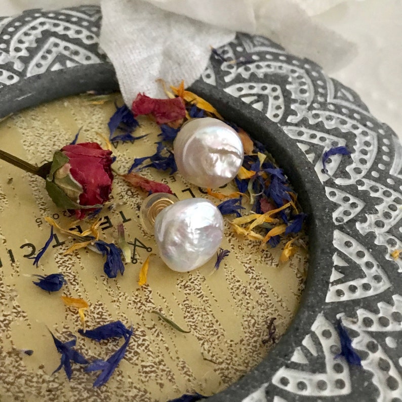May include: A pair of white pearl earrings with gold posts. The pearls are round and have an iridescent sheen. The earrings are displayed on a decorative surface with dried flowers.