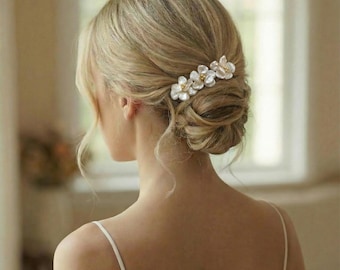 May include: A woman's blonde hair is styled in an updo, adorned with a delicate floral hair clip. The clip features three white flowers with gold-colored centers. The image is a close-up, focusing on the back of the head and the hair accessory.