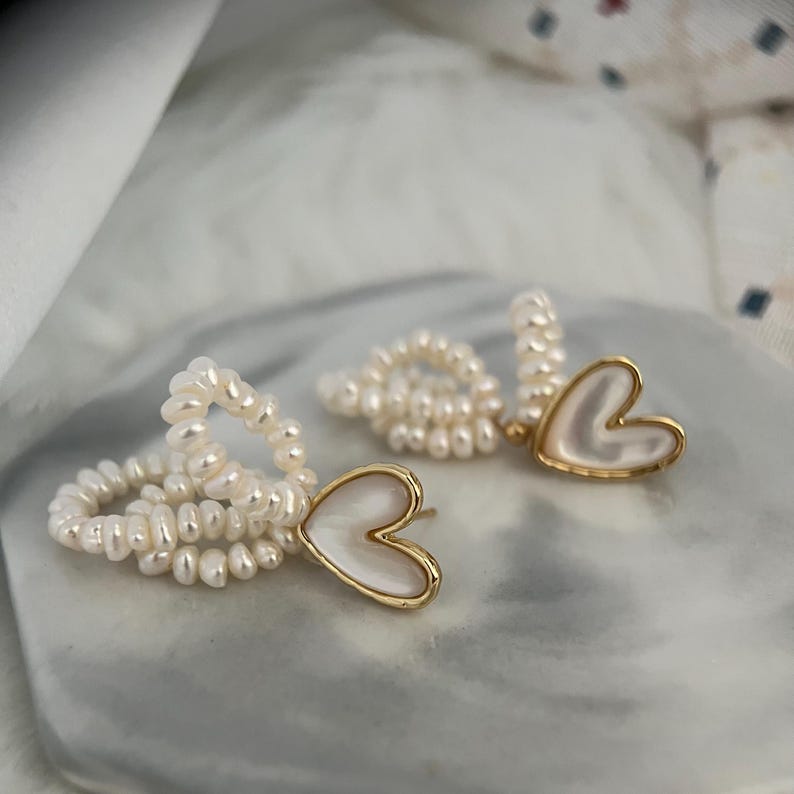 May include: A pair of pearl earrings with a heart-shaped pendant. The earrings are made of small white pearls and have a gold-tone finish.