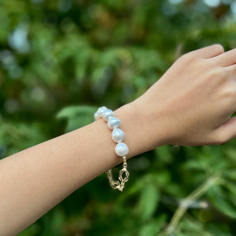 Puede incluir: Un brazalete de perlas blancas con cierre de cadena dorada. Las perlas son grandes e irregulares.