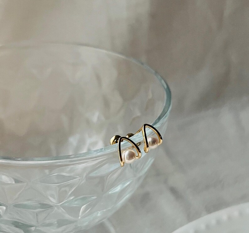 K&ouml;nnte beinhalten: Ein Paar goldfarbene Ohrringe mit jeweils einer kleinen Perle. Die Ohrringe sind dreieckig mit abgerundeter Oberseite.