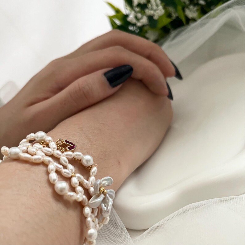May include: A multi-strand pearl bracelet with a gold clasp and a small red heart-shaped charm. The bracelet features irregular-shaped pearls and is worn on a wrist with black nail polish.