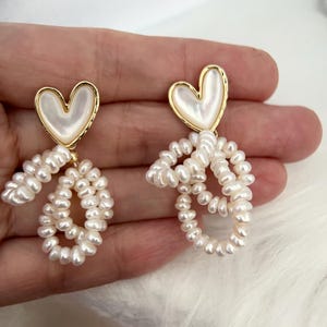 May include: A pair of gold heart-shaped earrings with a pearl-like finish. The earrings have a dangling design with multiple strands of small white pearls.