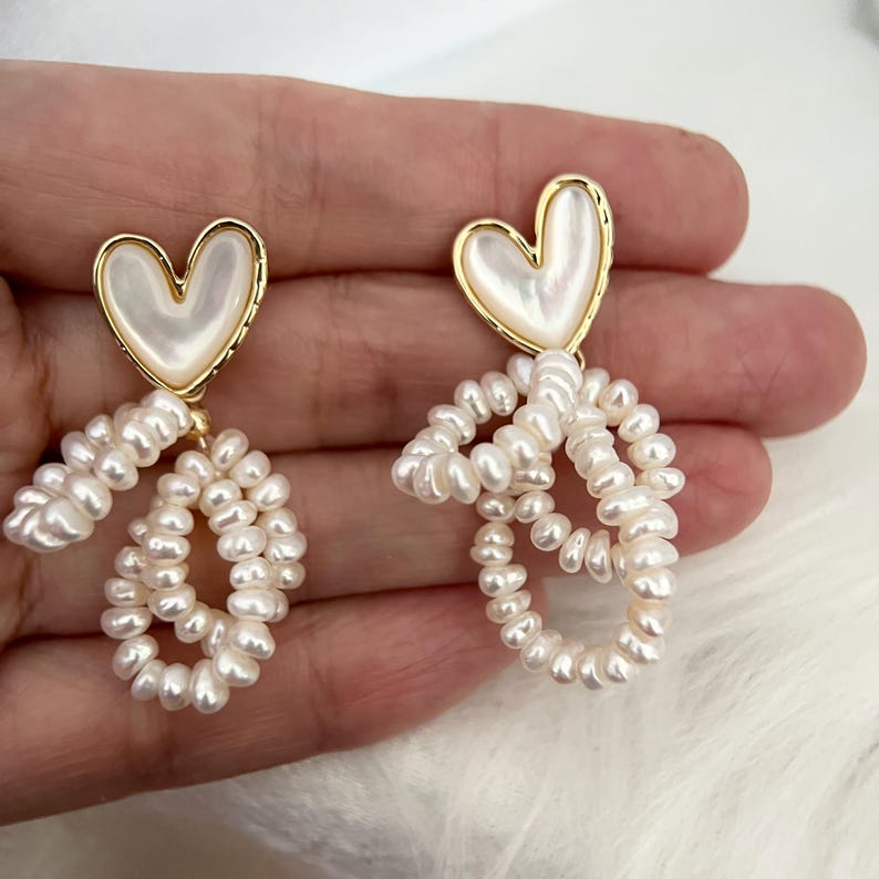May include: A pair of gold heart-shaped earrings with a pearl-like finish. The earrings have a dangling design with multiple strands of small white pearls.