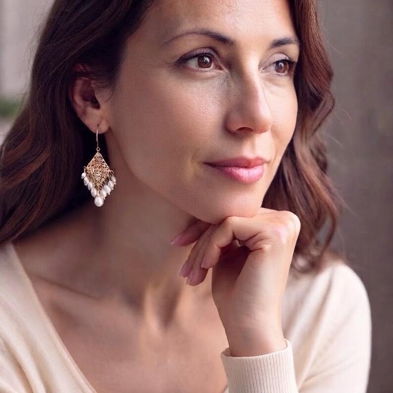 Peut inclure: Une boucle d'oreille pendante dor&eacute;e compos&eacute;e d'une pi&egrave;ce en m&eacute;tal filigran&eacute; en forme de losange suspendue &agrave; un crochet. Le bord inf&eacute;rieur du filigrane est orn&eacute; d'une s&eacute;rie de petites perles blanches en forme de goutte suspendues librement.