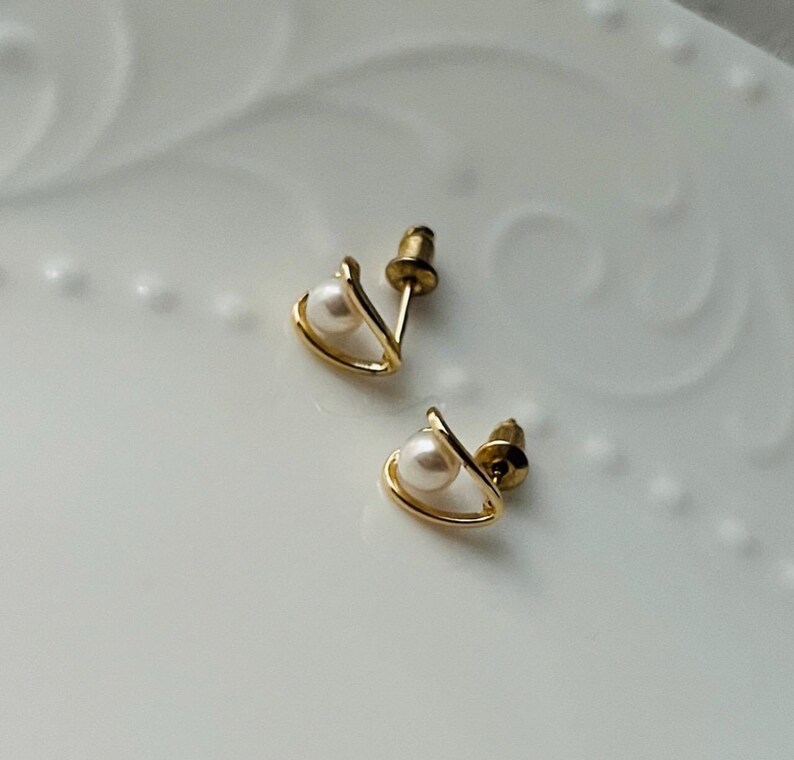 K&ouml;nnte beinhalten: Ein Paar goldene Ohrringe mit jeweils einer kleinen Perle. Die Ohrringe haben die Form eines Dreiecks mit einer abgerundeten Oberseite.