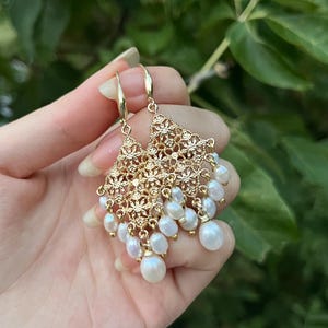 Peut inclure: Boucles d'oreilles pendantes dor&eacute;es avec un motif en filigrane ajour&eacute; de forme triangulaire, orn&eacute;es de multiples petites perles d'eau douce blanches en forme de goutte suspendues au bord inf&eacute;rieur de la structure m&eacute;tallique.