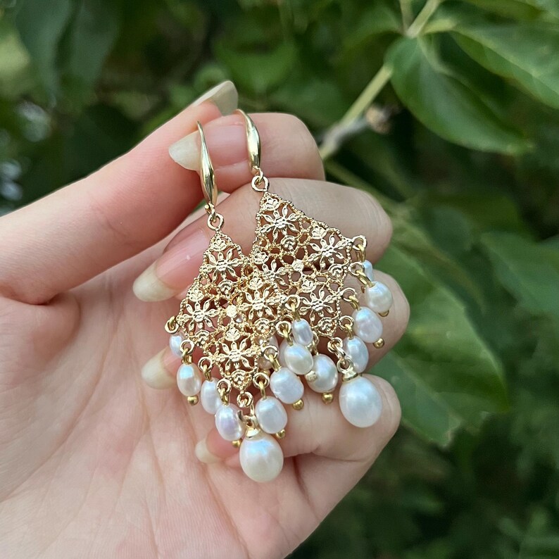 Peut inclure: Boucles d'oreilles pendantes dor&eacute;es avec un motif en filigrane ajour&eacute; de forme triangulaire, orn&eacute;es de multiples petites perles d'eau douce blanches en forme de goutte suspendues au bord inf&eacute;rieur de la structure m&eacute;tallique.