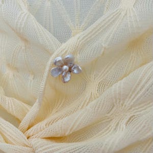 May include: Close-up of a cream-colored knitted cardigan with a floral brooch. The cardigan has a V-neck and a textured pattern. The brooch is made of multiple pearl-like petals.