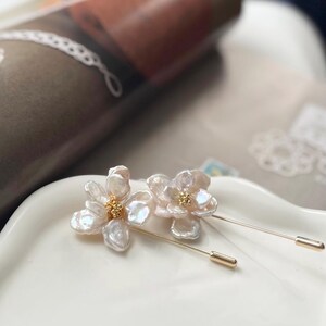 Puede incluir: Dos delicados broches de flores de perla con detalles dorados. Los broches est&aacute;n sobre una superficie blanca.