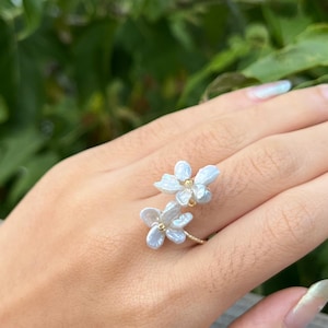 Puede incluir: Un delicado anillo de oro con dos acentos de flores de perla blanca. El anillo se lleva en el dedo de una persona.