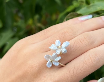 Puede incluir: Un delicado anillo de oro con dos acentos de flores de perla blanca. El anillo se lleva en el dedo de una persona.