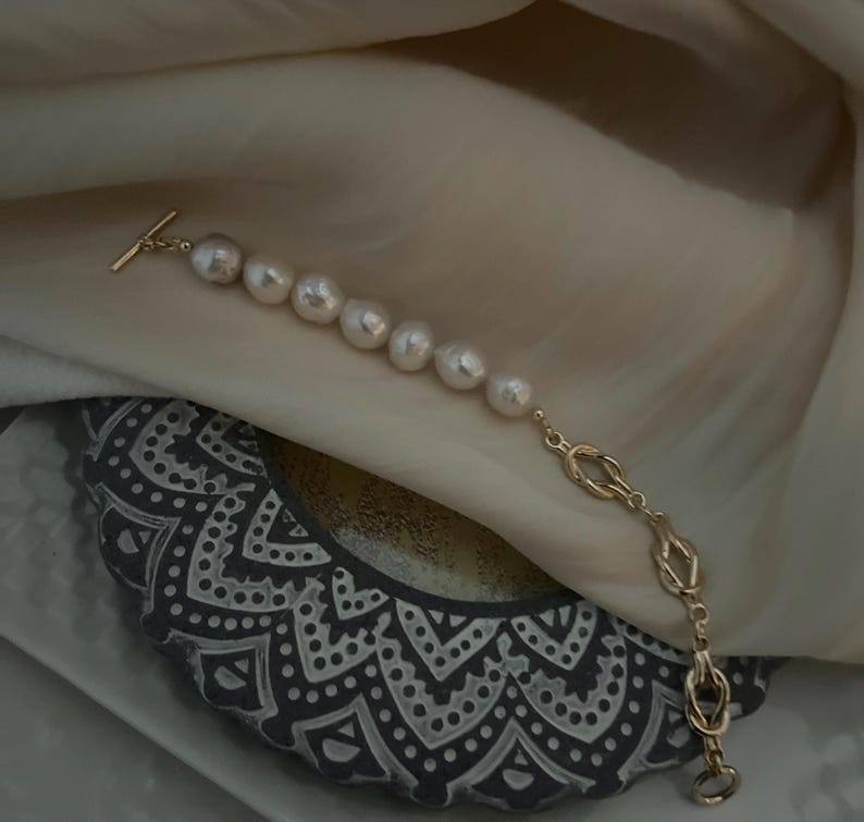 Puede incluir: Un brazalete de cadena dorada con una serie de perlas blancas grandes. La cadena tiene un dise&ntilde;o de nudo.