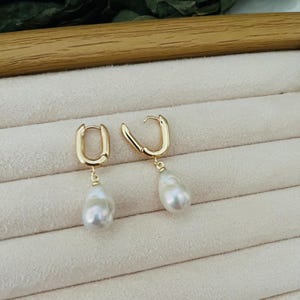 Peut inclure: Une paire de boucles d'oreilles créoles dorées avec un pendentif en perle blanche en forme de goutte. Les boucles d'oreilles sont présentées sur une surface beige clair. Design simple et élégant.