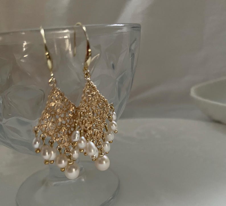 Peut inclure: Une paire de boucles d'oreilles pendantes dor&eacute;es compos&eacute;es d'un maillage m&eacute;tallique filigrane d&eacute;licat et complexe en forme de triangle, orn&eacute; de multiples petites perles d'eau douce blanches et rondes suspendues aux crochets.