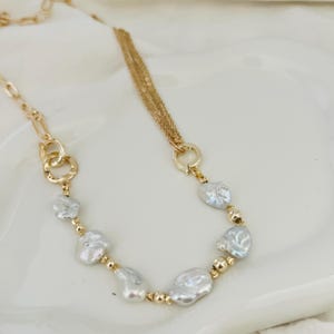 Peut inclure: Un collier dor&eacute; orn&eacute; de perles blanches irr&eacute;guli&egrave;res et de petites perles dor&eacute;es. Le collier pr&eacute;sente une cha&icirc;ne et un design &agrave; plusieurs brins, avec une fermeture &agrave; fermoir. Le bijou est pr&eacute;sent&eacute; sur une surface blanche.