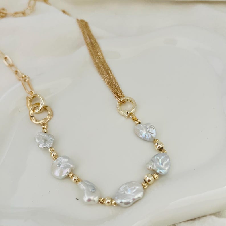Peut inclure: Un collier dor&eacute; orn&eacute; de perles blanches irr&eacute;guli&egrave;res et de petites perles dor&eacute;es. Le collier pr&eacute;sente une cha&icirc;ne et un design &agrave; plusieurs brins, avec une fermeture &agrave; fermoir. Le bijou est pr&eacute;sent&eacute; sur une surface blanche.