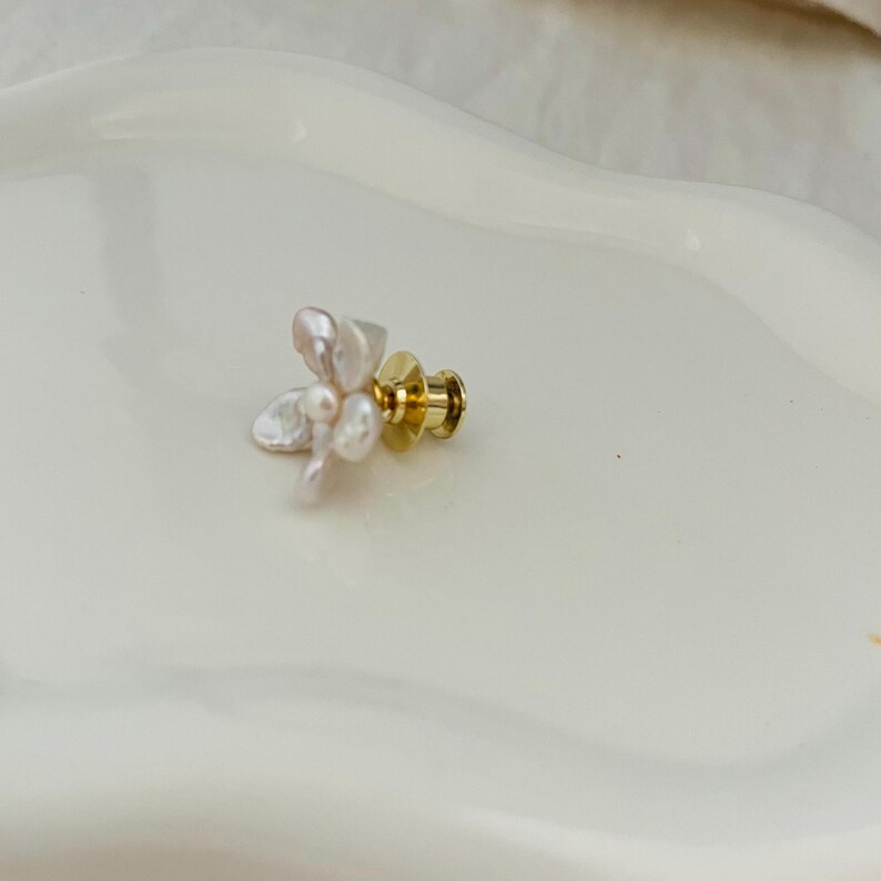 May include: A delicate floral pin featuring a cluster of off-white, irregularly shaped pearls. The pin has a gold-colored post and backing. The pearls are arranged to resemble a flower, creating an elegant accessory.