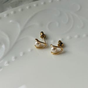 K&ouml;nnte beinhalten: Ein Paar goldene Ohrringe mit einer kleinen Perle in jedem. Die Ohrringe haben die Form einer geschwungenen Linie.