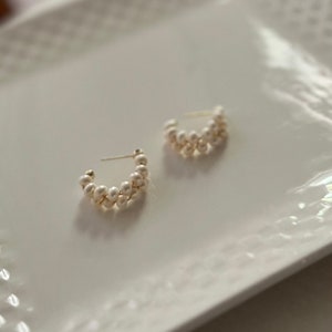 May include: A pair of gold hoop earrings with white pearl accents. The earrings are made of small, round pearls that are strung together in a continuous loop.