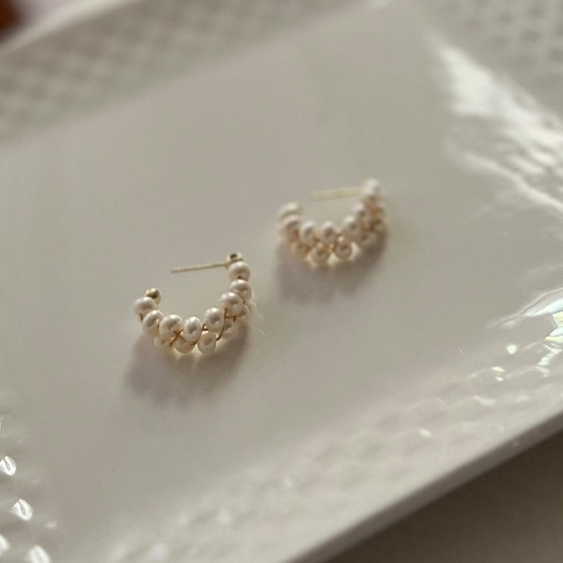May include: A pair of gold hoop earrings with white pearl accents. The earrings are made of small, round pearls that are strung together in a continuous loop.