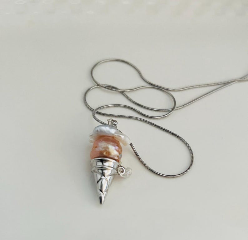 Peut inclure: Un collier en cha&icirc;ne argent avec un pendentif en forme de cornet de glace. Le cornet est rempli de trois couches de perles roses et blanches.
